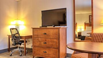 A hotel room with a wooden desk, a chair, a television on a cabinet, a round dining table, and a mirror.