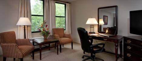 A hotel room with a desk, two chairs, a laptop, a TV, and a large window with curtains.
