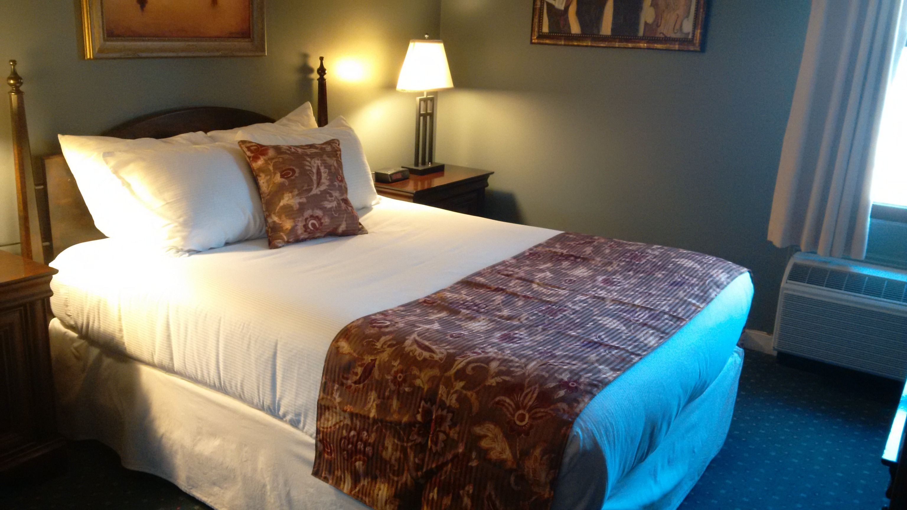 A neatly made bed with white linens and a patterned throw blanket, a bedside lamp, and a framed picture on the wall.