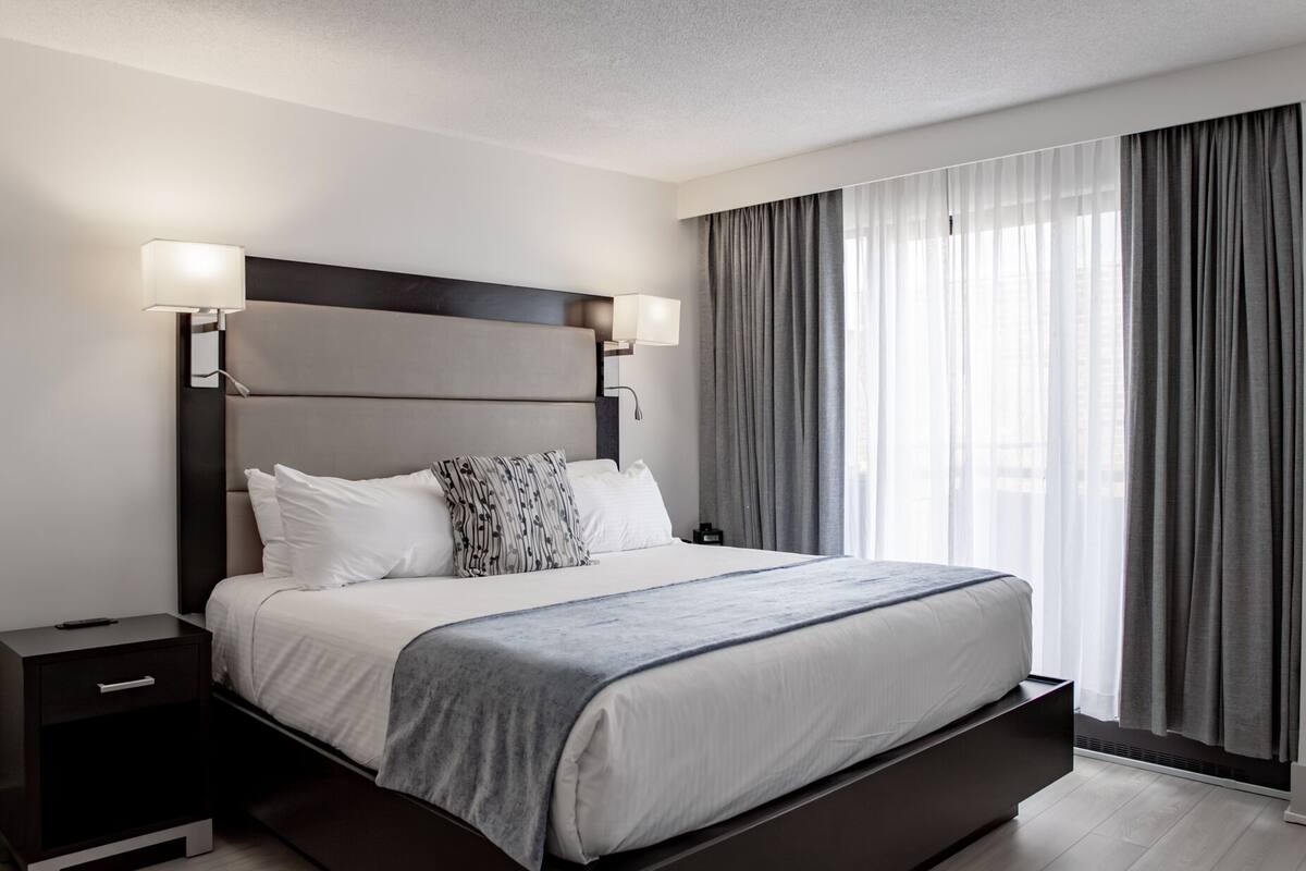 A modern hotel room with a large bed, two bedside lamps, a nightstand, and a view of the city through sheer curtains.