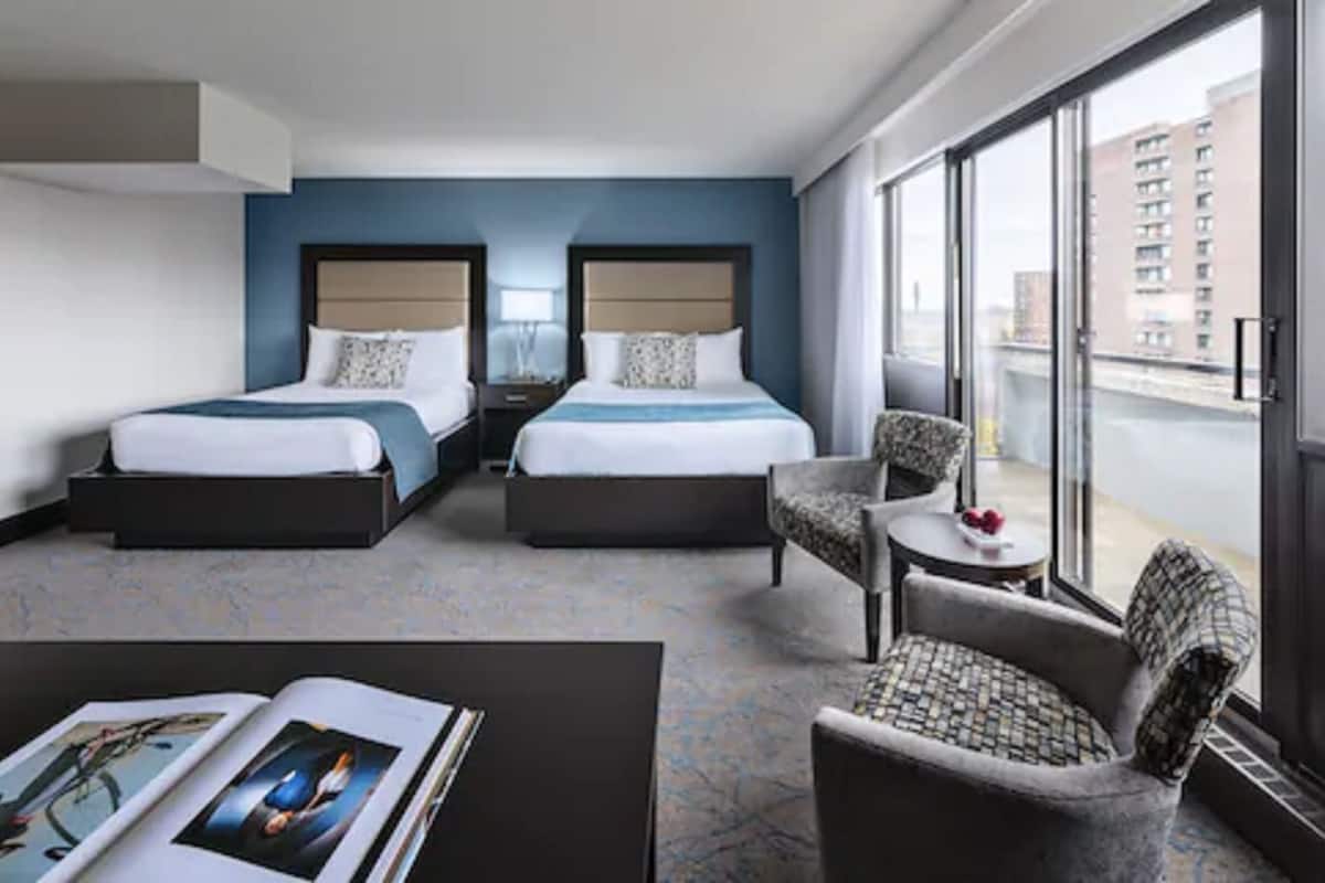 A hotel room with two beds, a small table, a chair, and a large window offering a city view.