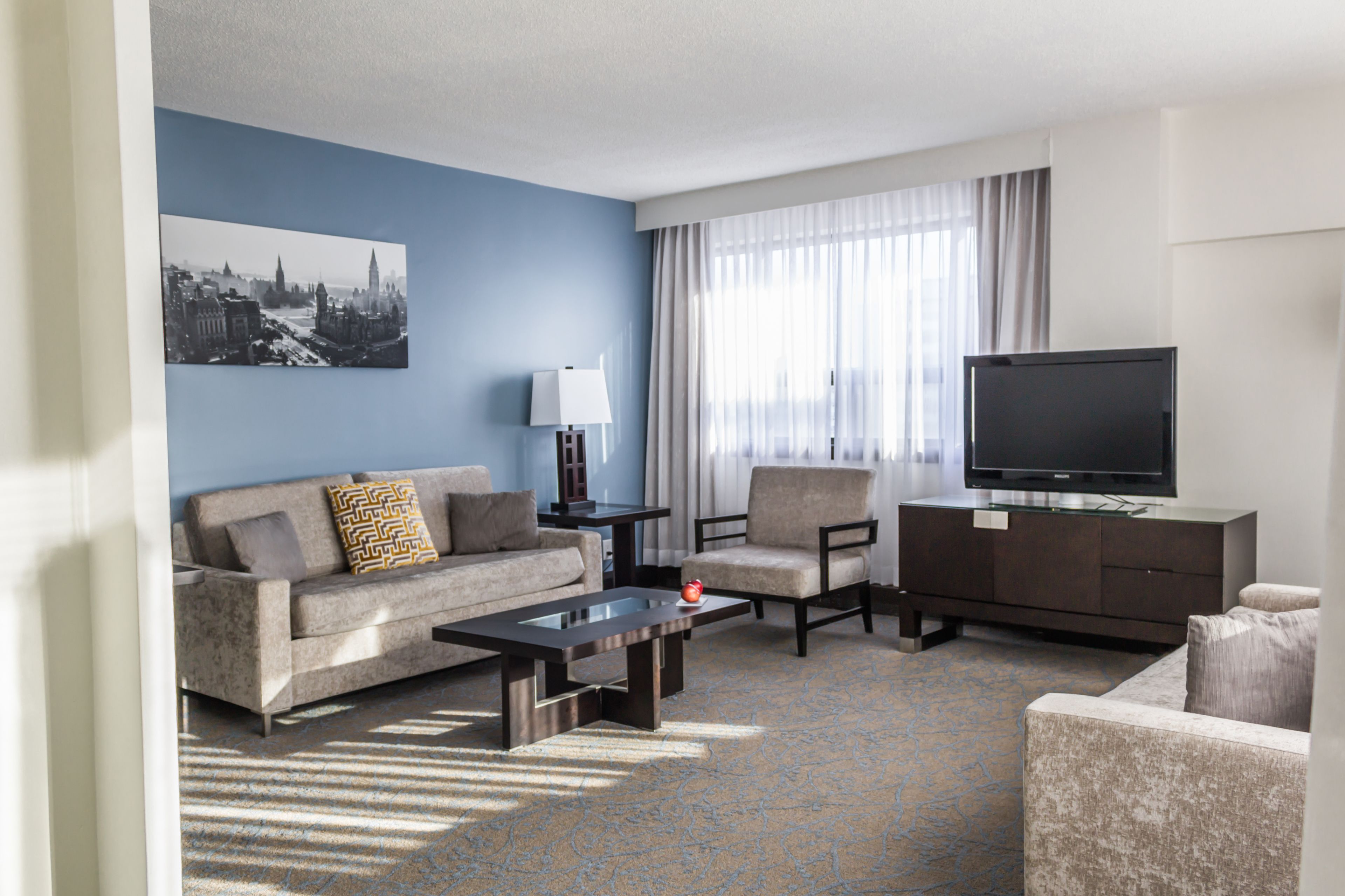 A modern hotel room with a blue accent wall, a large flat-screen TV, a sofa, a chair, a coffee table, and a lamp.