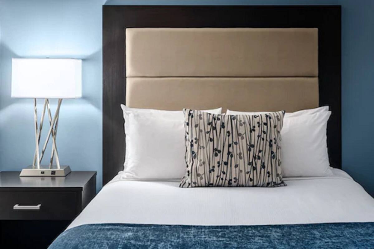 A neatly made bed with a headboard, a bedside table with a lamp, and a wall with a blue hue.