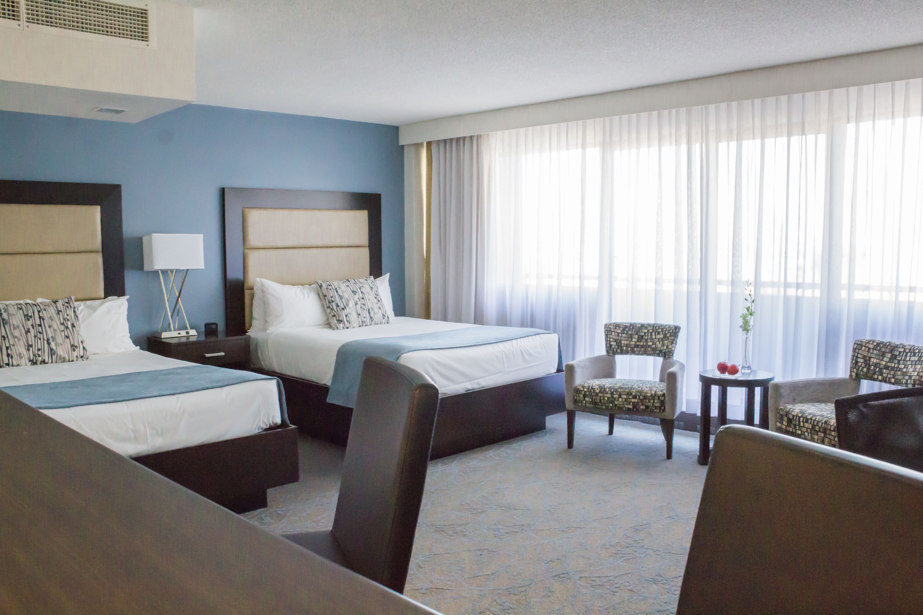 A hotel room with two beds, a sitting area with a chair and a small table, and a large window with curtains.