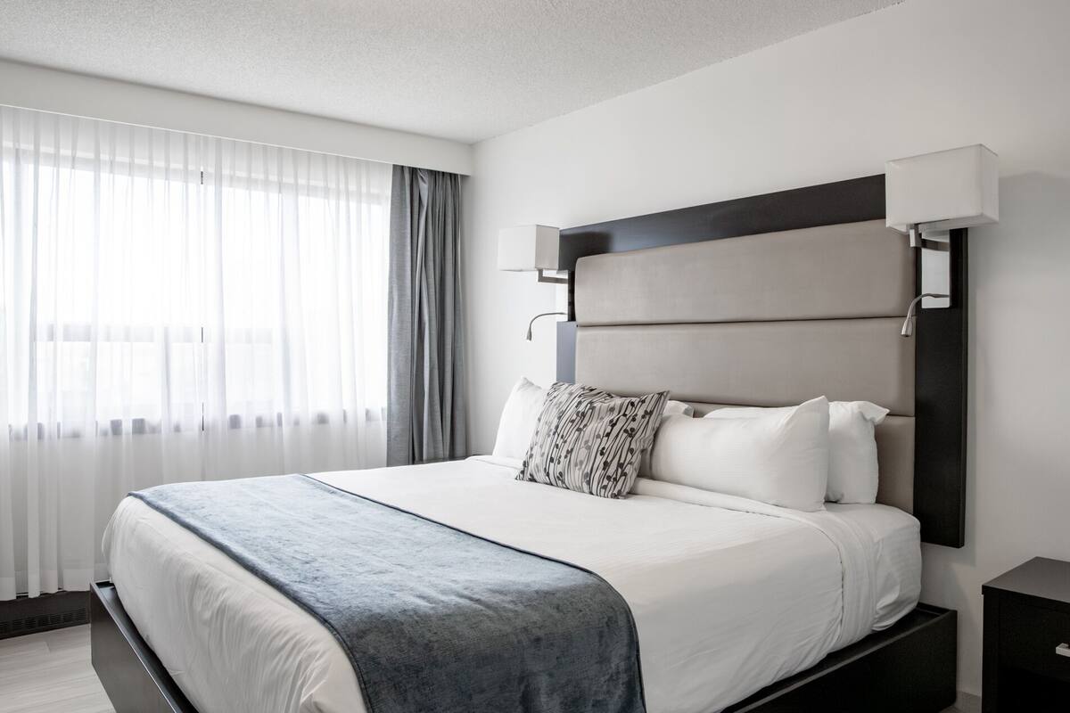 A modern hotel room with a large bed, a bedside table, a nightstand, and a window with sheer curtains.