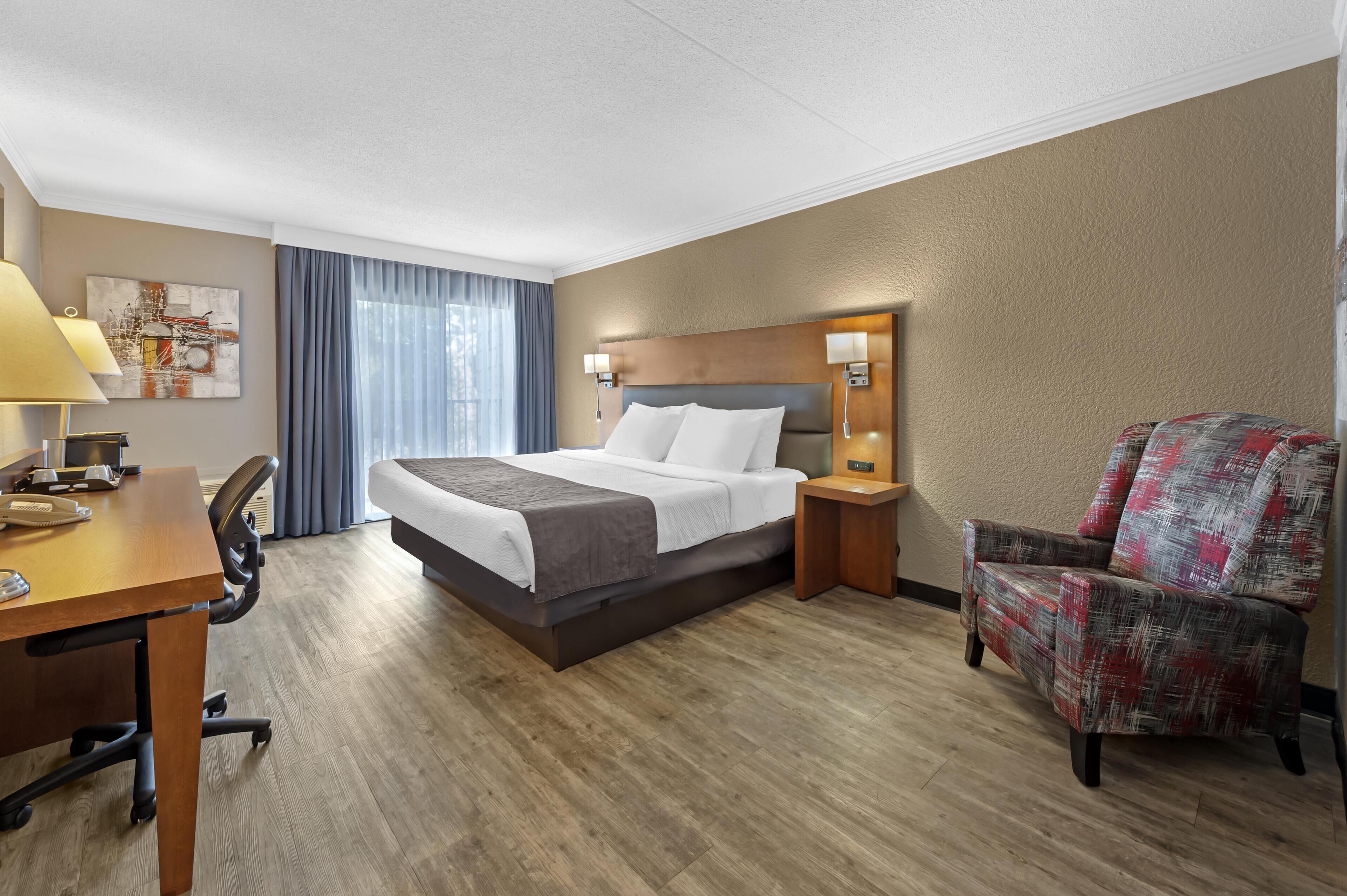 A hotel room with a bed, desk, chair, and a nightstand with a lamp.