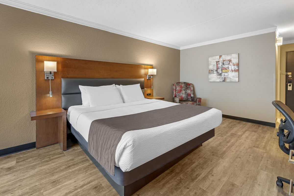 Una habitación de hotel con una cama grande, cabecera de madera, lámparas de noche, una silla y un cuadro en la pared.