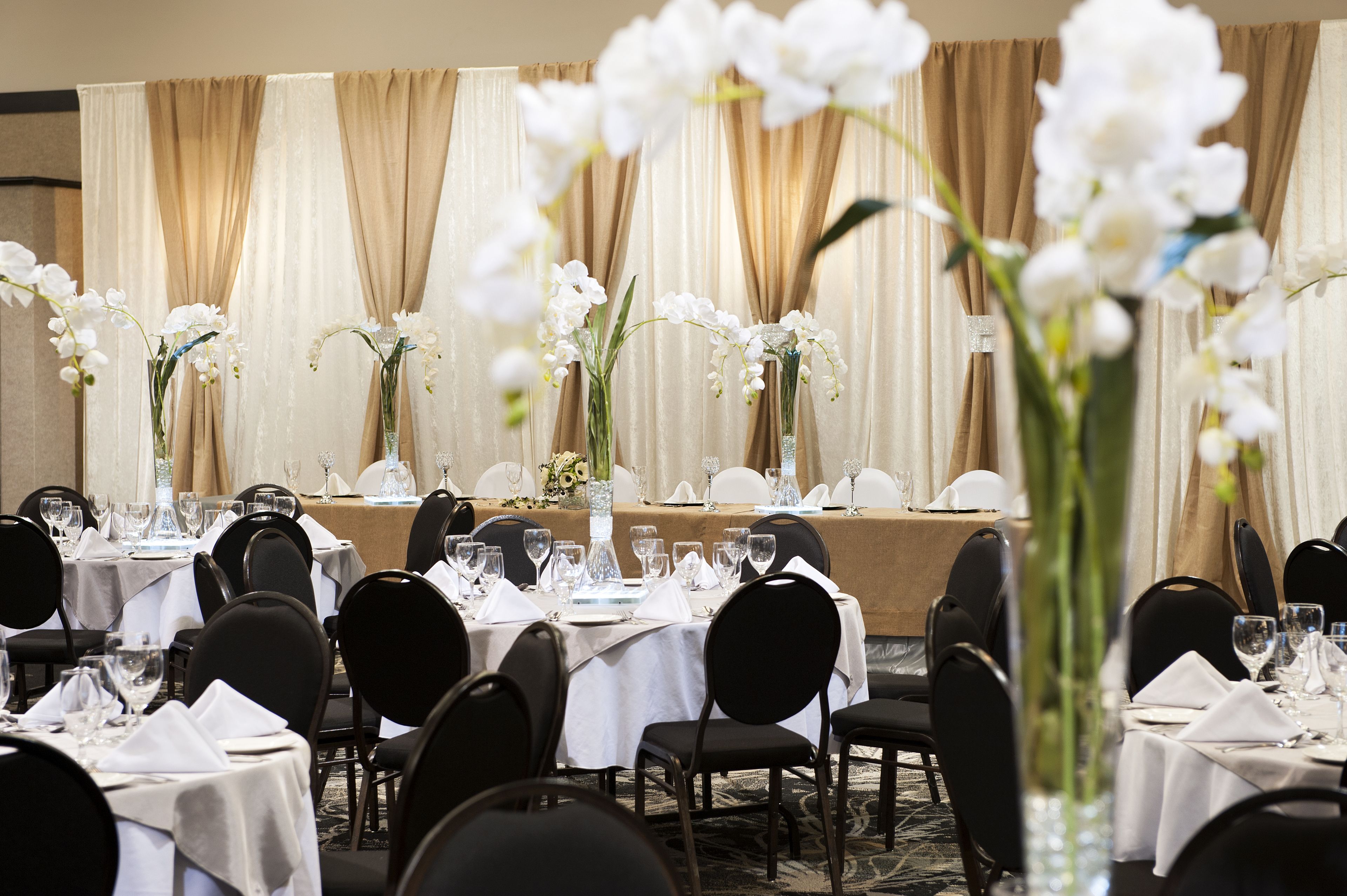 Een feestzaal met ronde tafels gedekt voor een diner, elk met een witte tafelkleed en zwarte stoelen.