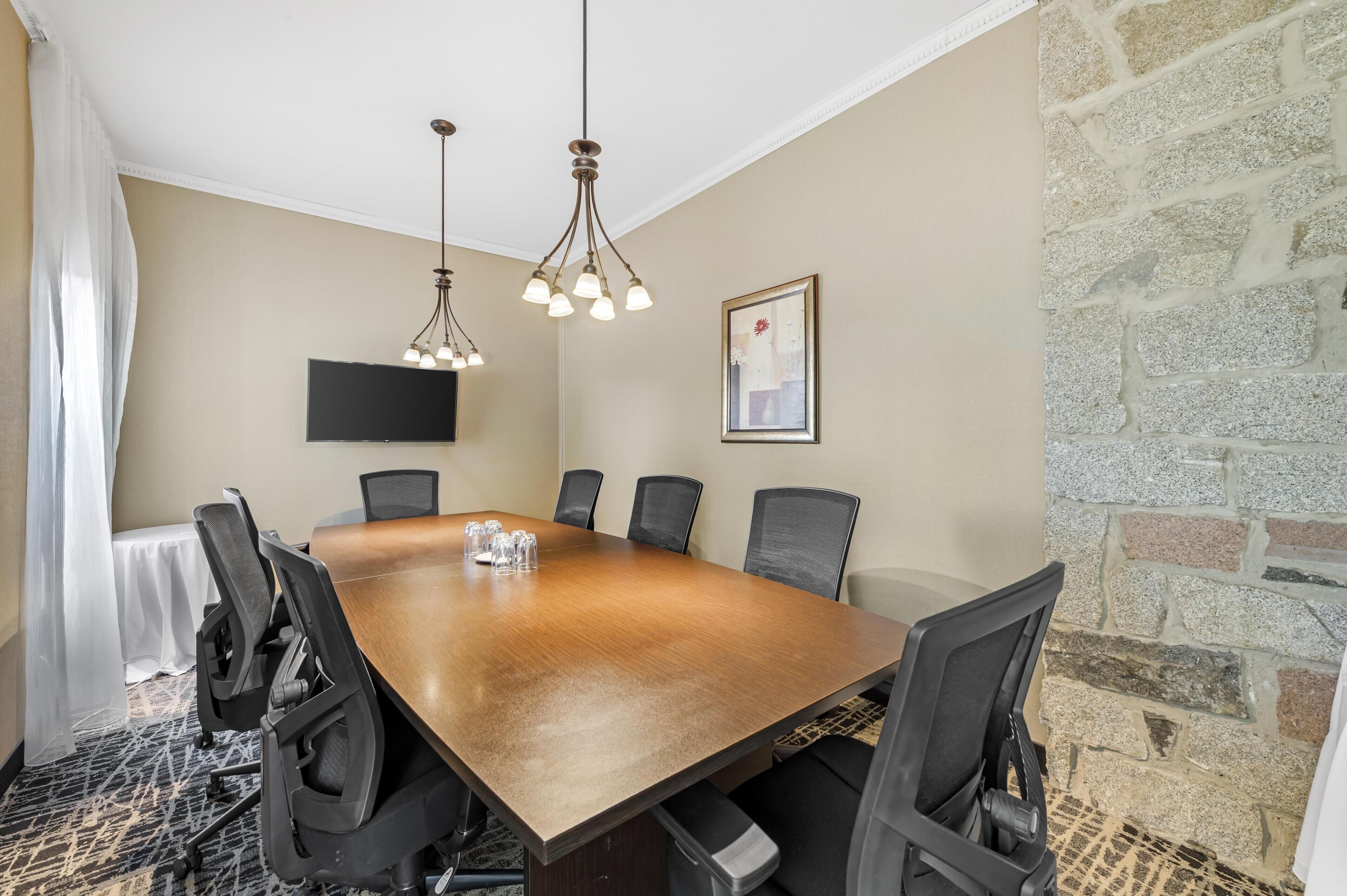 A conference room with a large wooden table, black chairs, a mounted TV, and a stone wall.
