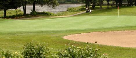 Campo de golf con un cuerpo de agua al fondo, rodeado de árboles y una zona arenosa.