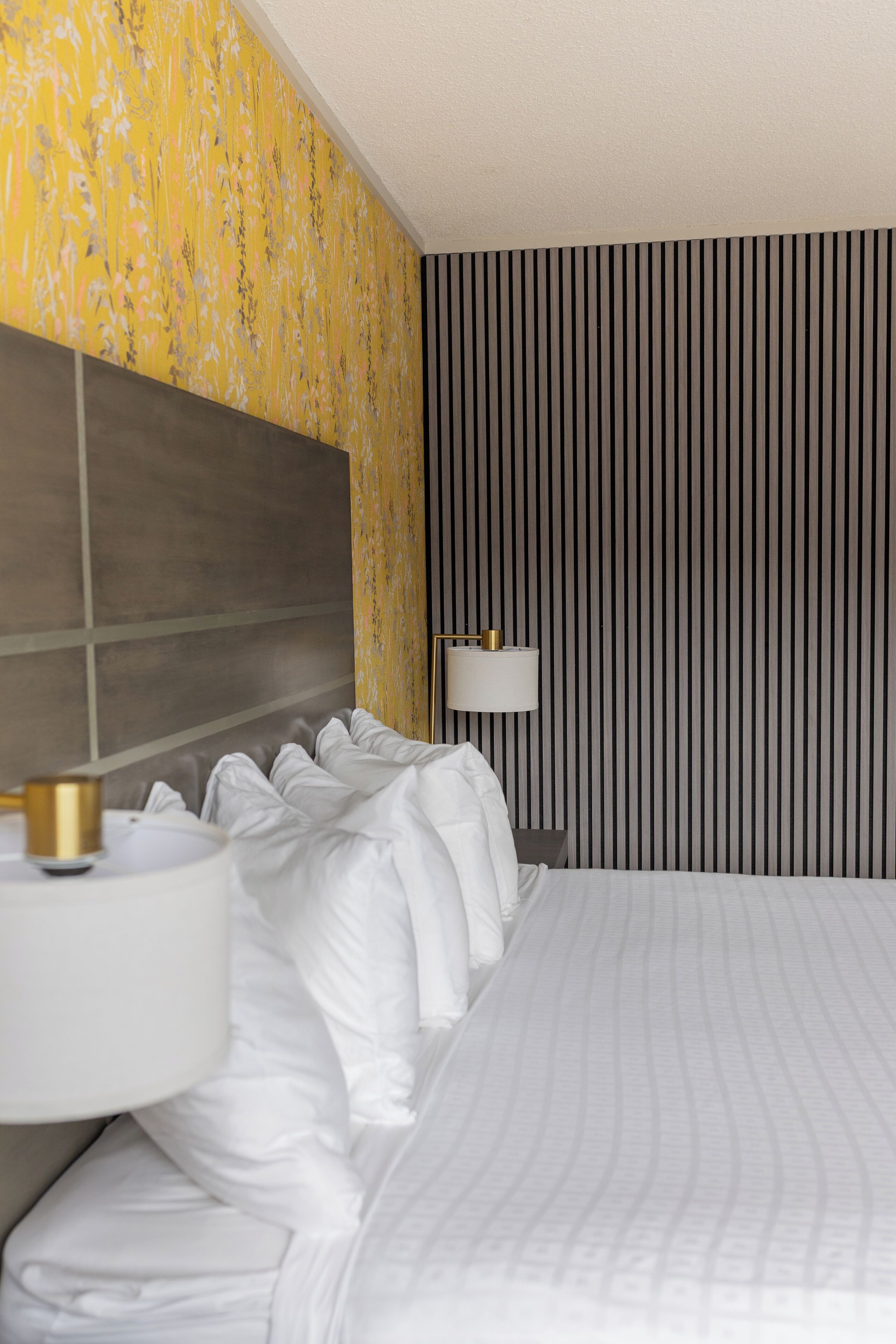 Una habitació d'hotel amb un llit gran, una làmpada de nit i una paret amb ratlles verticals.