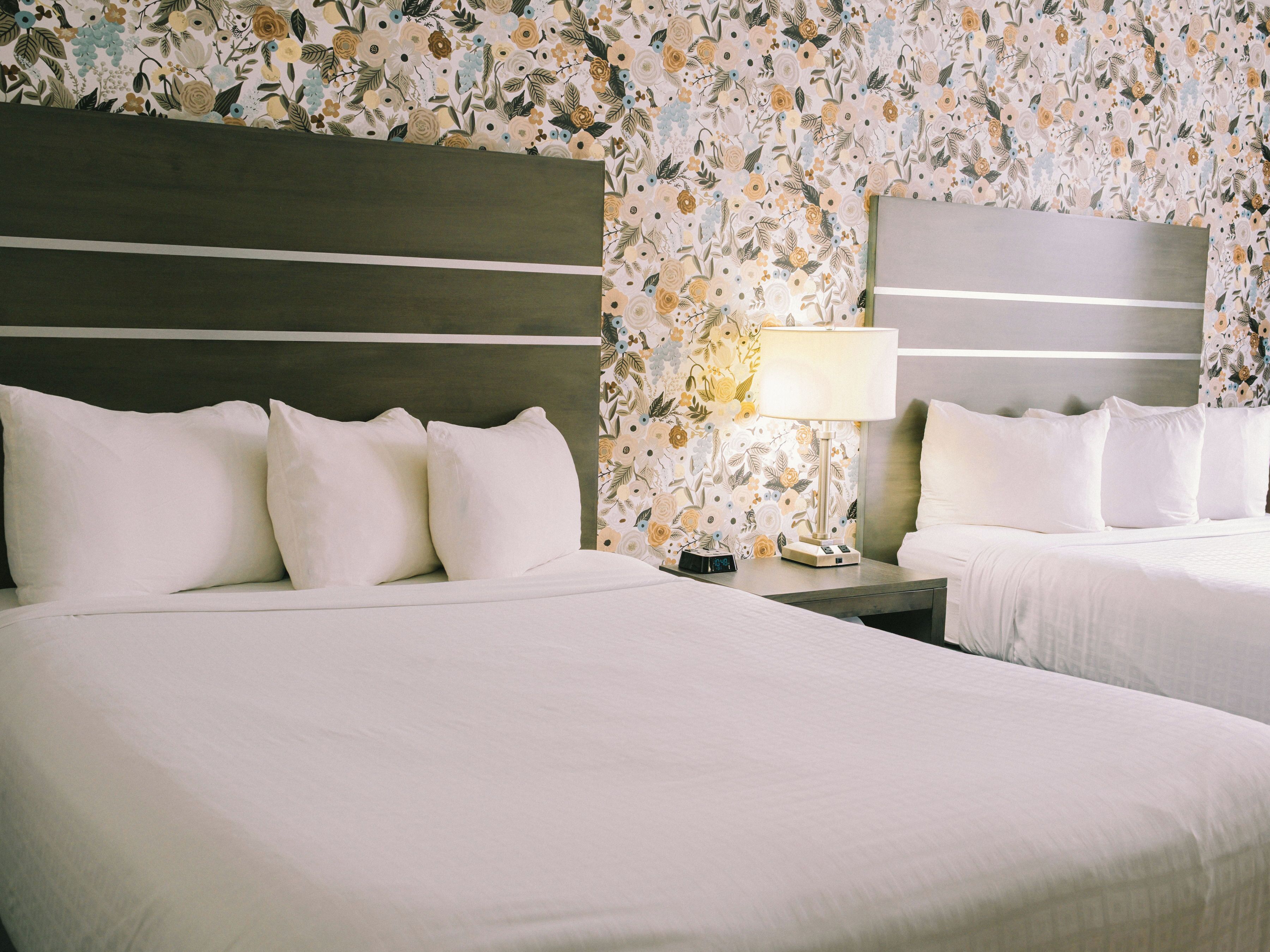 A hotel room with two beds, a nightstand with a lamp, and floral wallpaper.