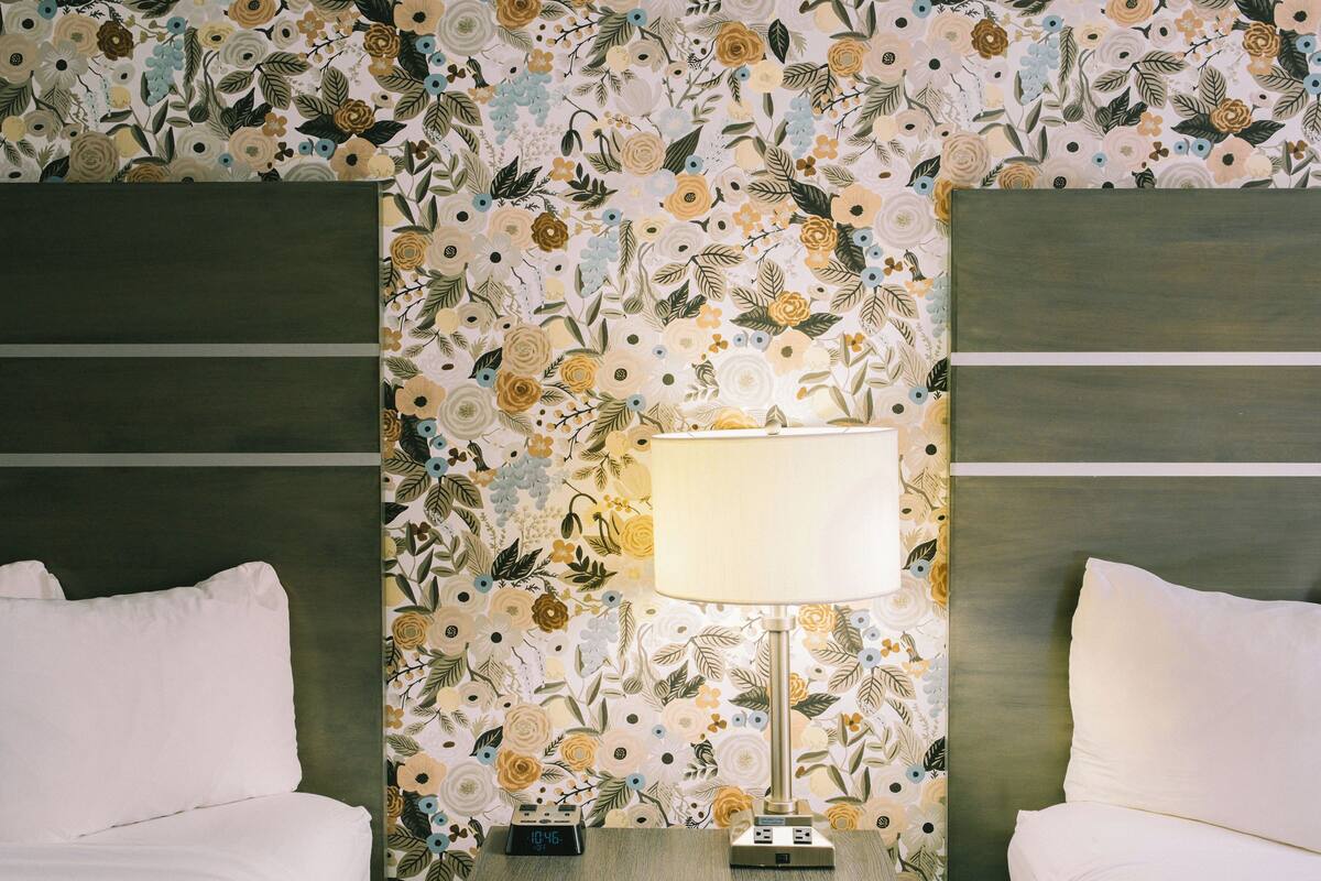 Una habitación de hotel con papel tapiz floral, una mesita de noche con lámpara y dos camas.