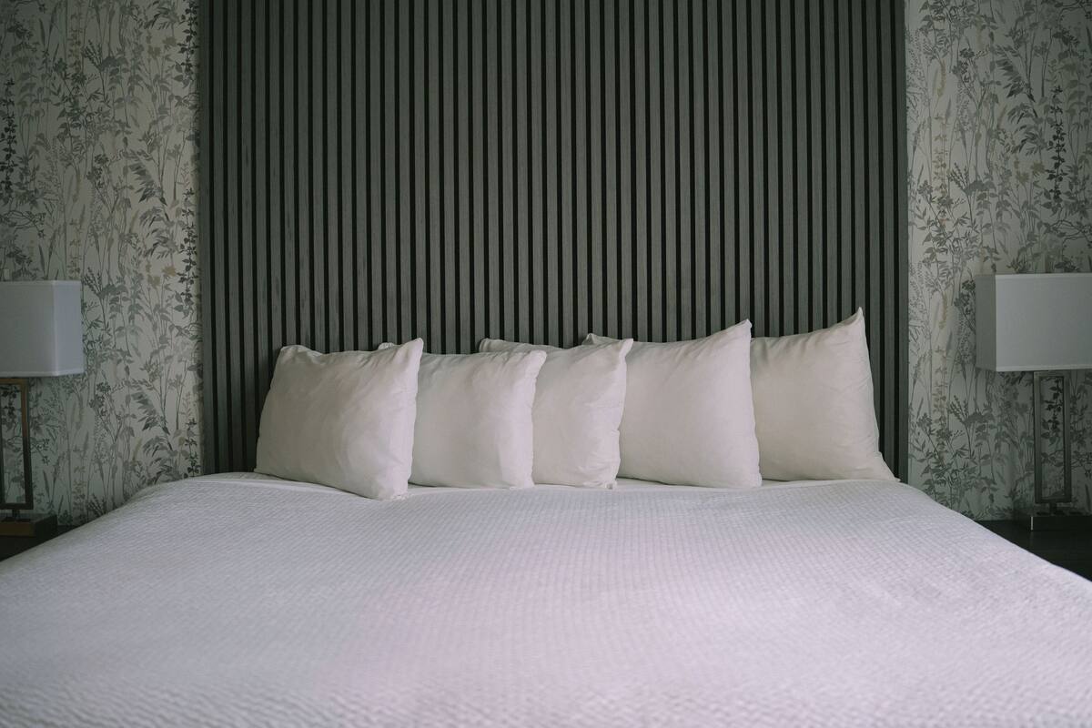 Una cama impecablemente tendida con un edredón blanco texturizado, un cabecero a rayas verticales y papel tapiz floral en las paredes.