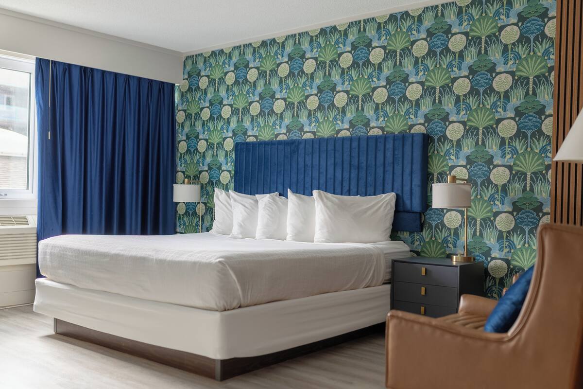 Una cama bien hecha con ropa blanca, un cabecero azul y una mesita de noche con una lámpara. La habitación tiene un papel tapiz estampado y una ventana con cortinas azules.
