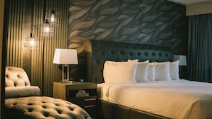 Una habitación de hotel con un cabecero capitonado, una cama grande con sábanas blancas, un sillón capitonado, una mesita de noche con lámpara y una pared con un diseño ondulado.