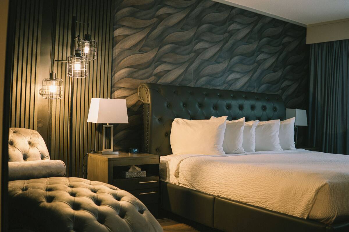 Una habitación de hotel con un cabecero capitonado, una cama grande con sábanas blancas, un sillón capitonado, una mesita de noche con lámpara y una pared con un diseño ondulado.