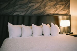 Una cama bien hecha con ropa blanca y almohadas, una pared con textura y una mesita de noche con lámpara y control remoto.