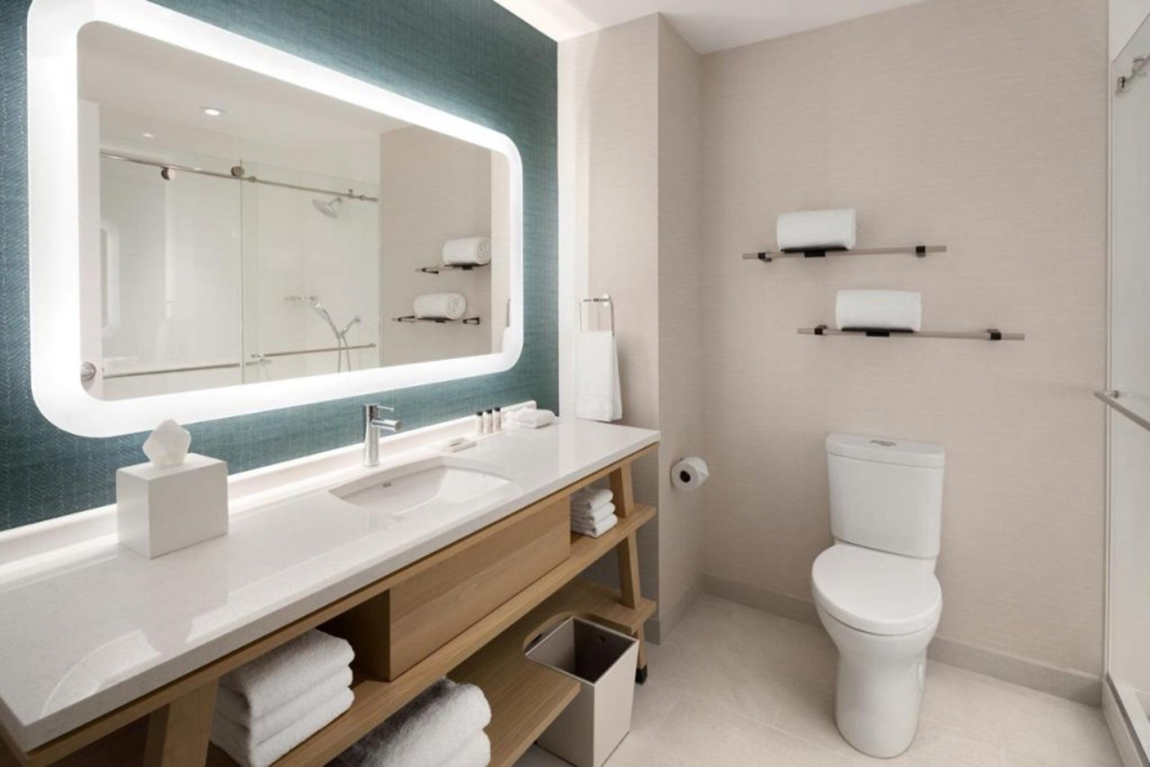 A modern bathroom with a large mirror, white sink, and toilet. There are shelves with towels and toiletries.
