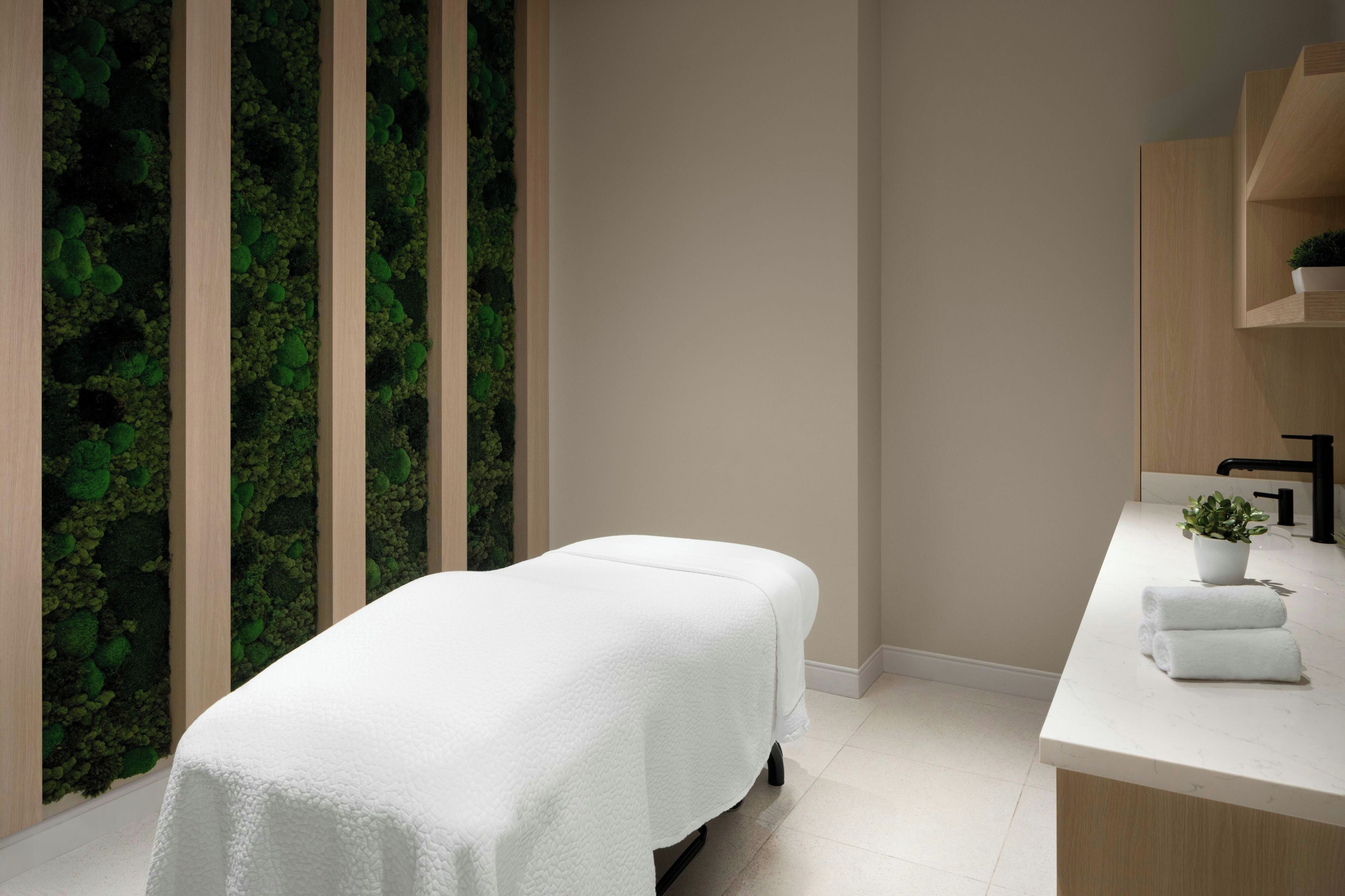A modern spa room with a massage table, a wooden wall with a green moss-like texture, and a white countertop with a sink and a potted plant.