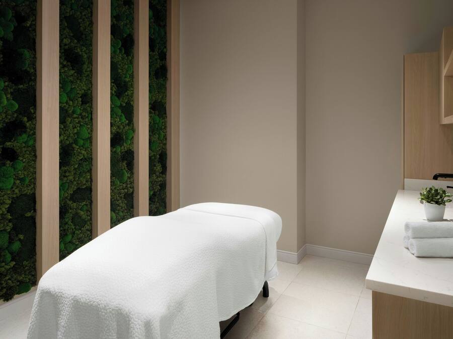 A modern spa room with a massage table, a wooden wall with a green moss-like texture, and a white countertop with a sink and a potted plant.