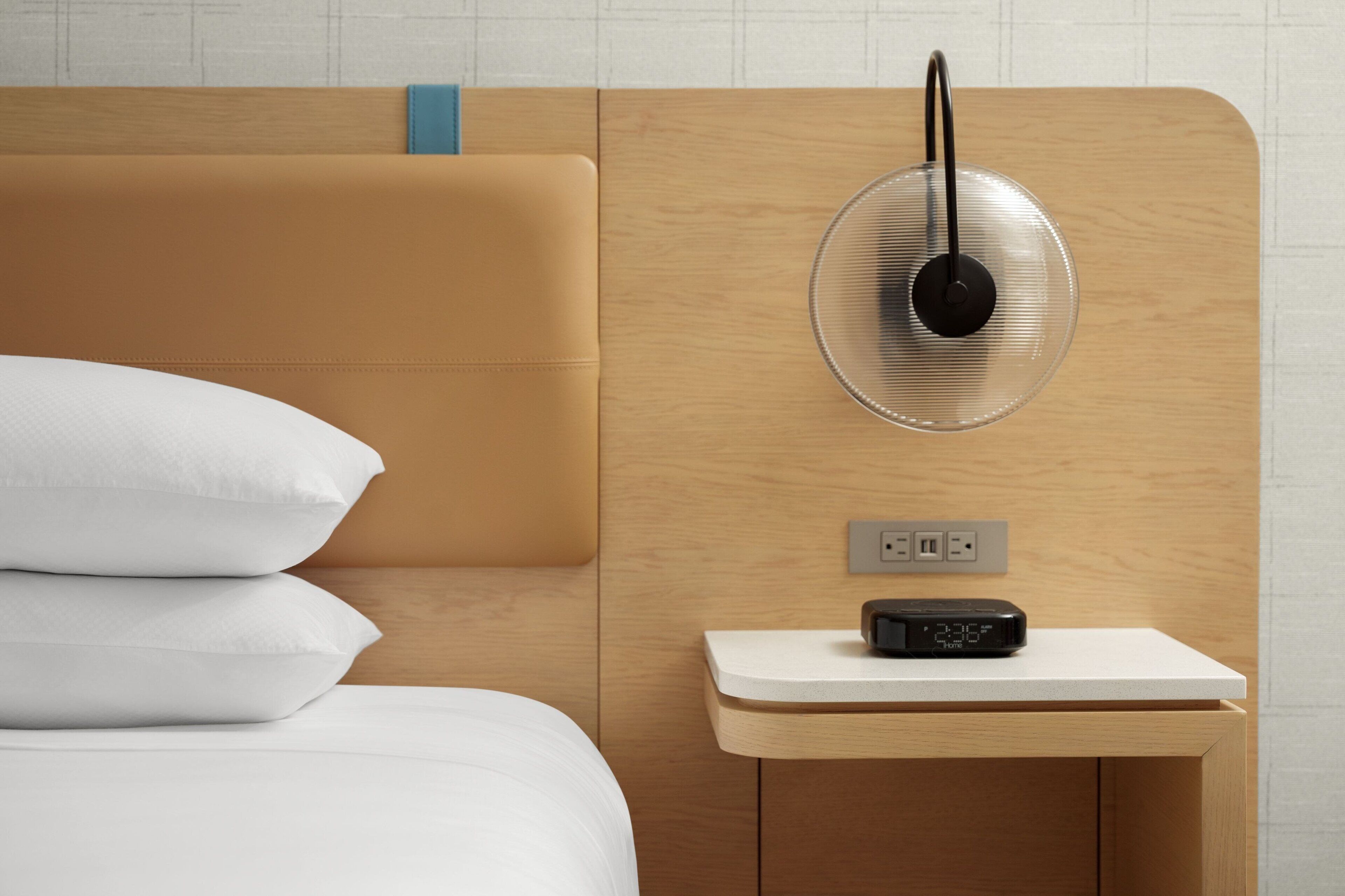 A neatly made bed with white linens and pillows, a bedside table with a clock, and a wall-mounted fan.