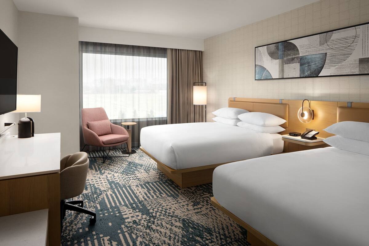 Quarto de hotel com duas camas, uma escrivaninha, uma cadeira, uma televisão e uma janela com cortinas.