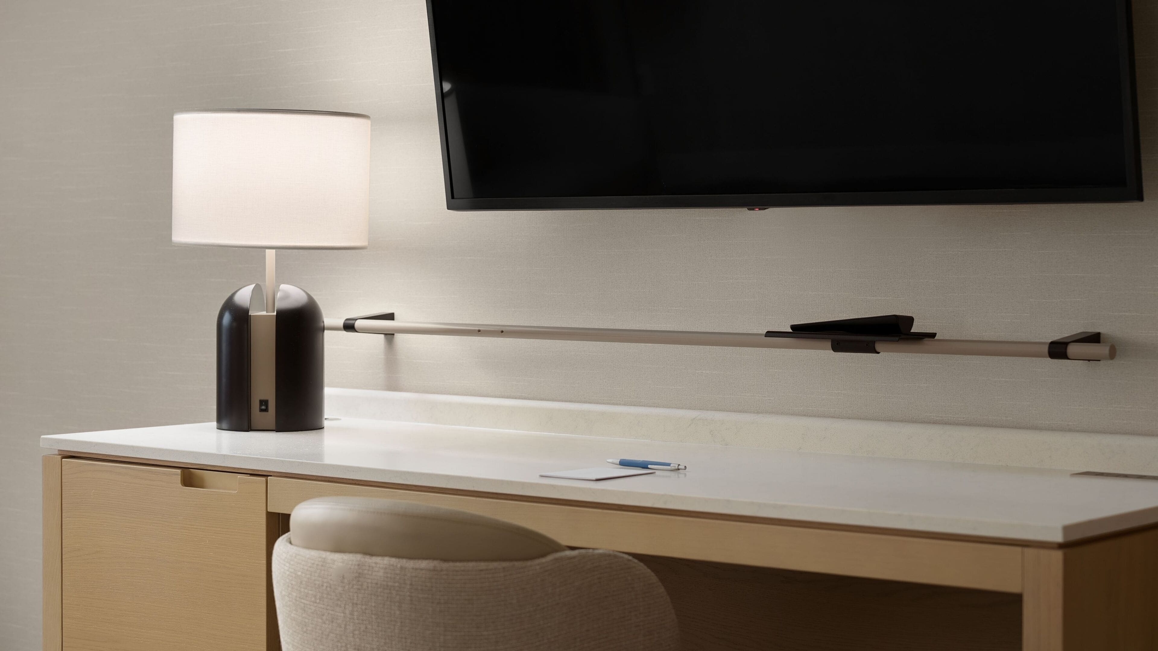 A hotel room with a desk, a lamp, a flat-screen TV mounted on the wall, and a chair.