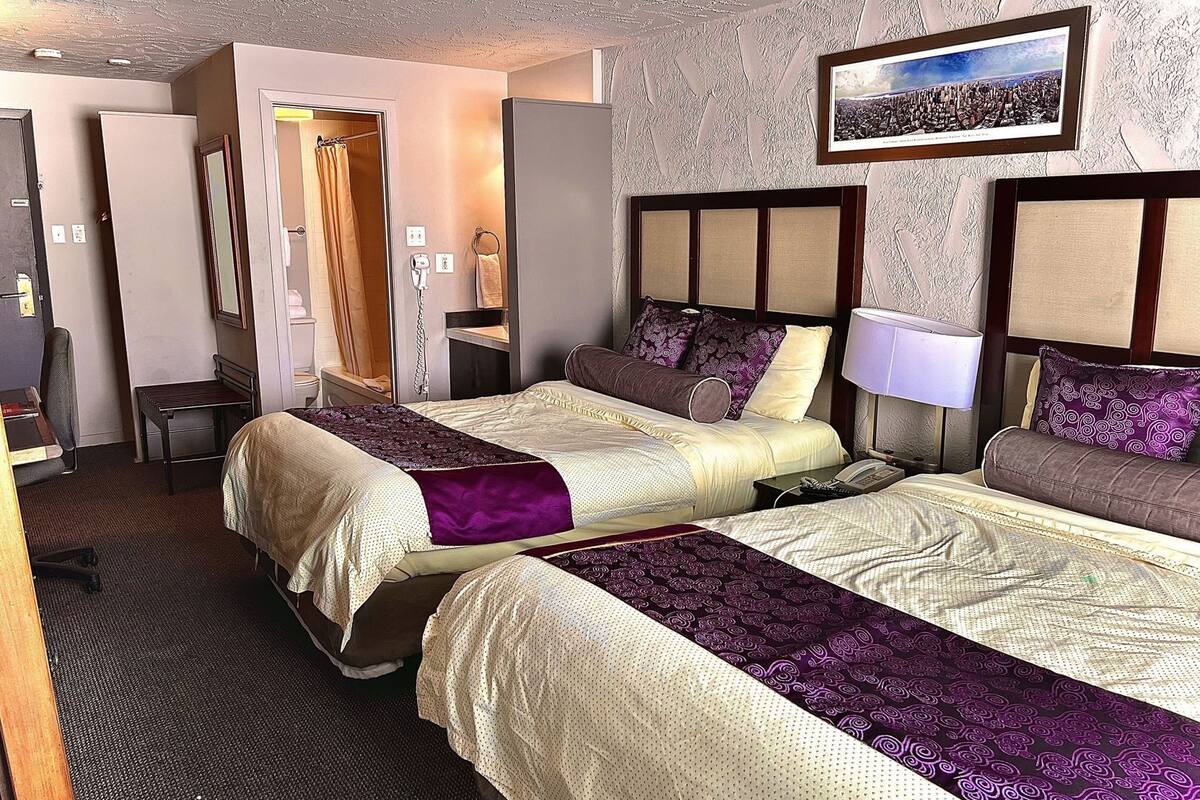 A hotel room with two beds, a desk, a chair, a bathroom, and a framed cityscape picture on the wall.