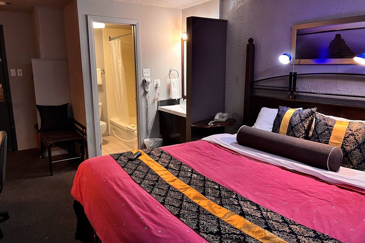 A hotel room with a bed, a chair, a desk with a phone, a mirror, and a shower area.