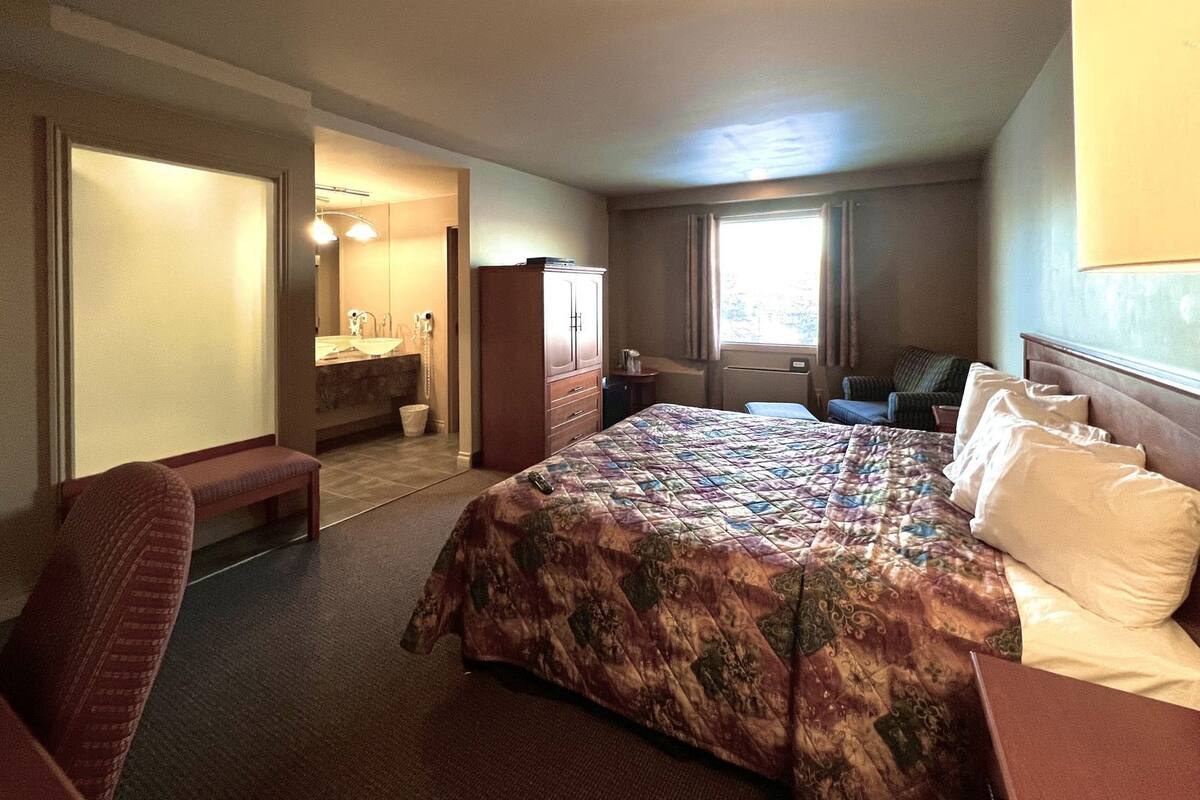 A hotel room with a large bed, a desk, a chair, a bench, a bathroom with a sink and mirror, and a window with curtains.