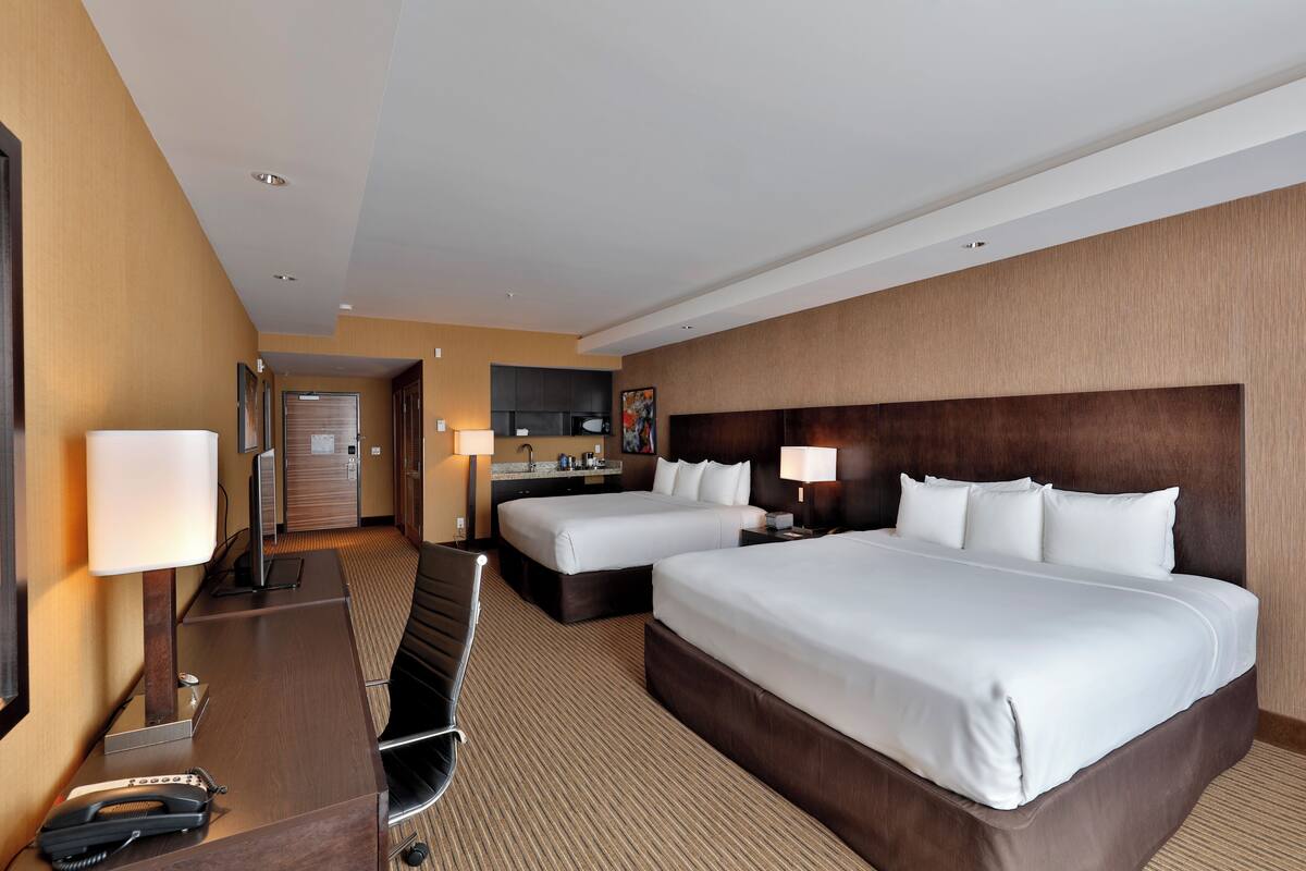 A hotel room with two beds, a desk with a telephone, a chair, a desk lamp, and a small kitchenette.