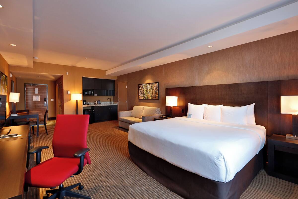 A hotel room with a large bed, a desk with a chair, a sofa, and a kitchenette area.