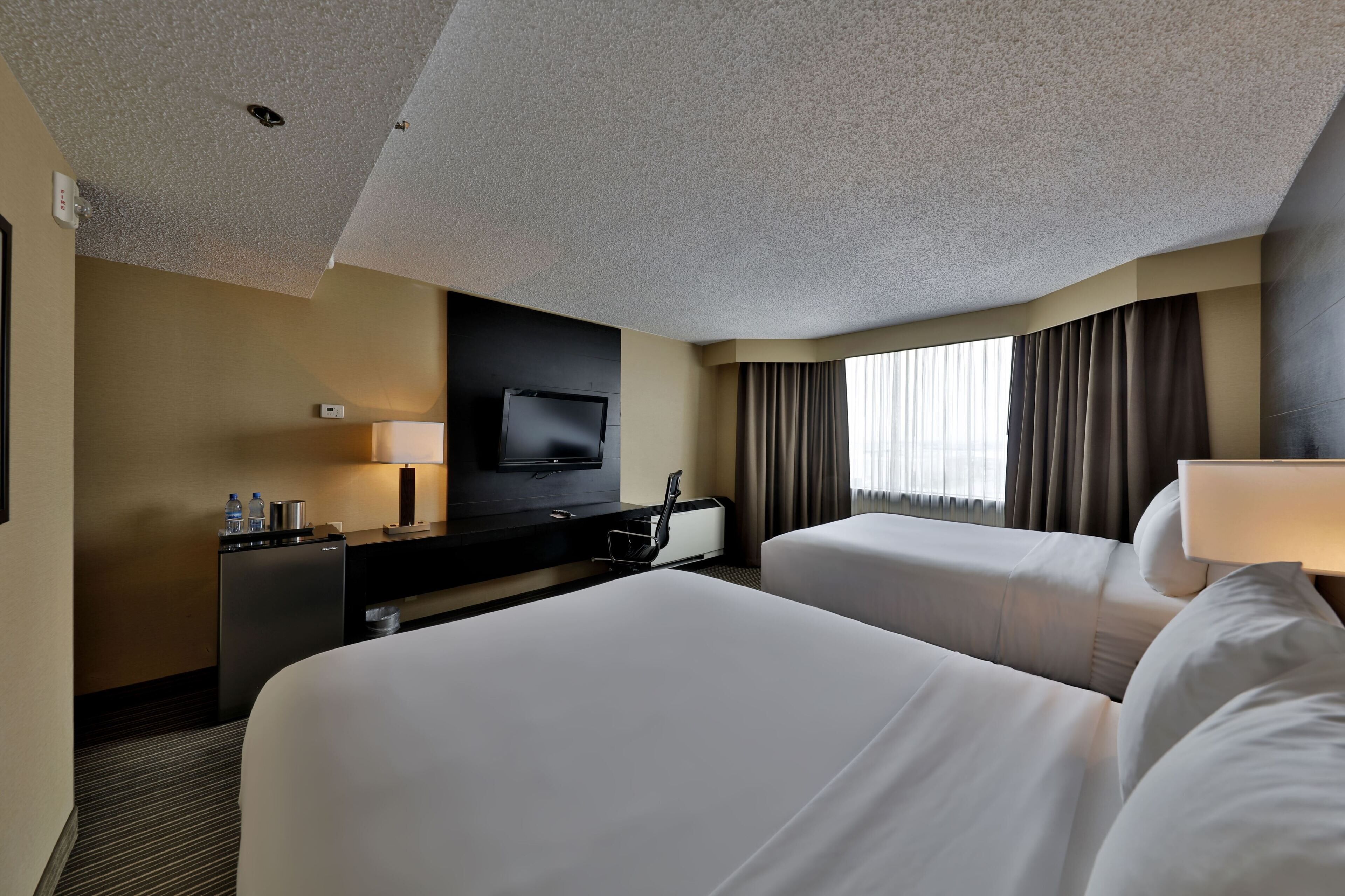 A hotel room with two beds, a TV, a desk with a lamp, and a window with curtains.