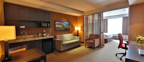 A hotel room with a kitchenette, a sofa, a desk, and a chair.