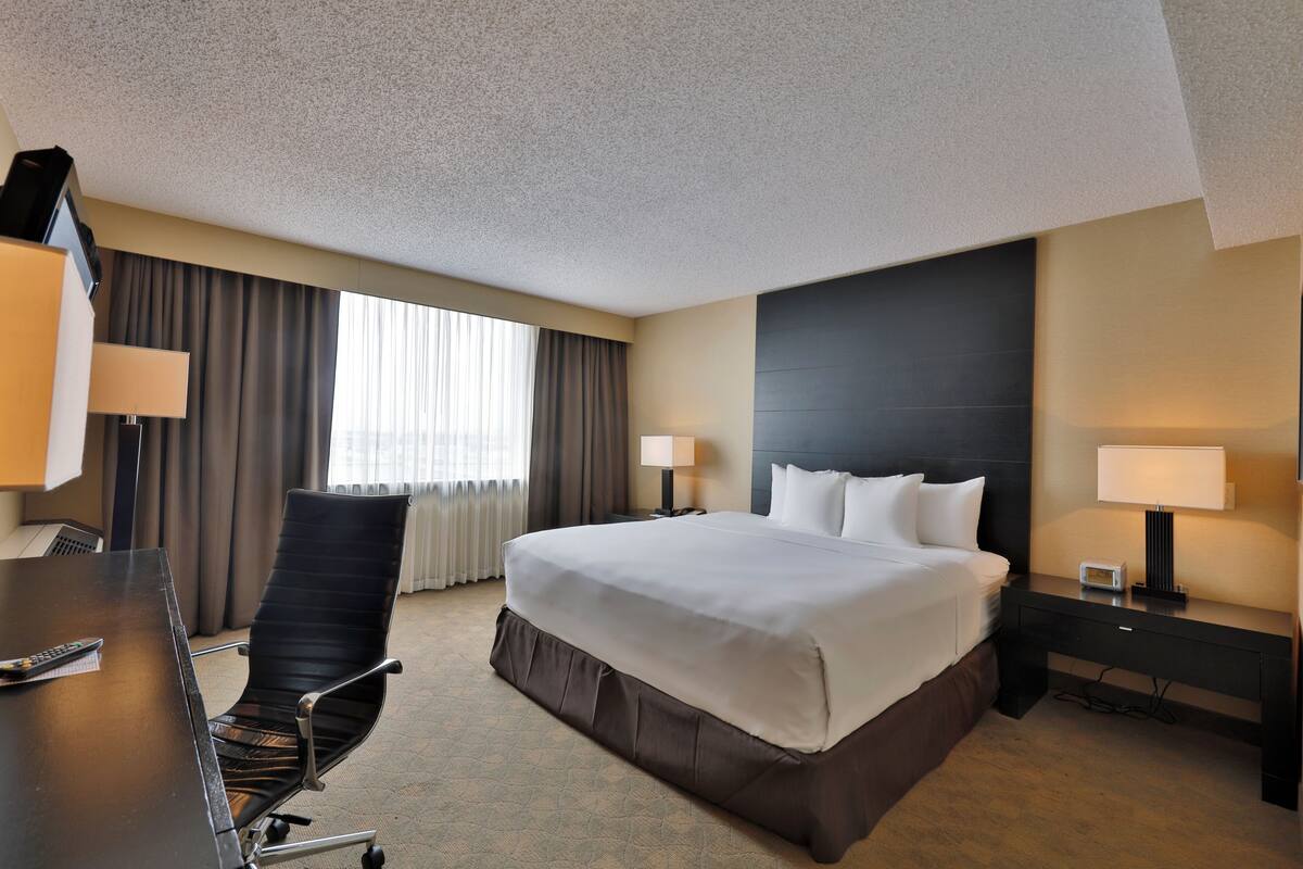 A hotel room with a large bed, a desk with a chair, two bedside lamps, and a window with curtains.