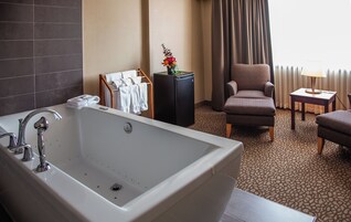 Un baño de hotel con una bañera grande, un perchero con toallas, una mesita de noche con una lámpara y una silla.