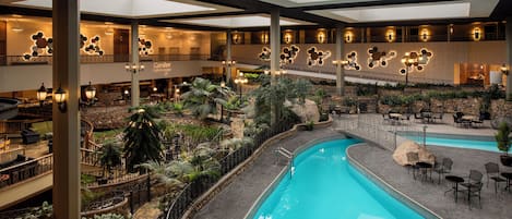 Un área de piscina interior con varios niveles, con una piscina central rodeada de zonas de estar y vegetación.