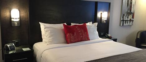 Habitación de hotel con una cama grande, ropa de cama blanca, un cojín rojo decorativo, lámparas de noche, una mesita de noche con un teléfono y un cuadro en la pared.