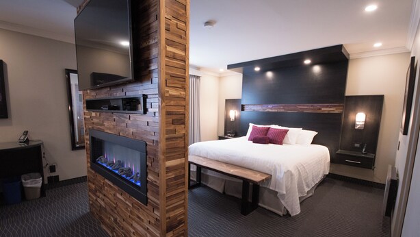 A modern hotel room with a fireplace, a bed with white bedding, a bench, and a flat-screen TV mounted on a wooden wall. - Hôtel des Eskers (Amos)