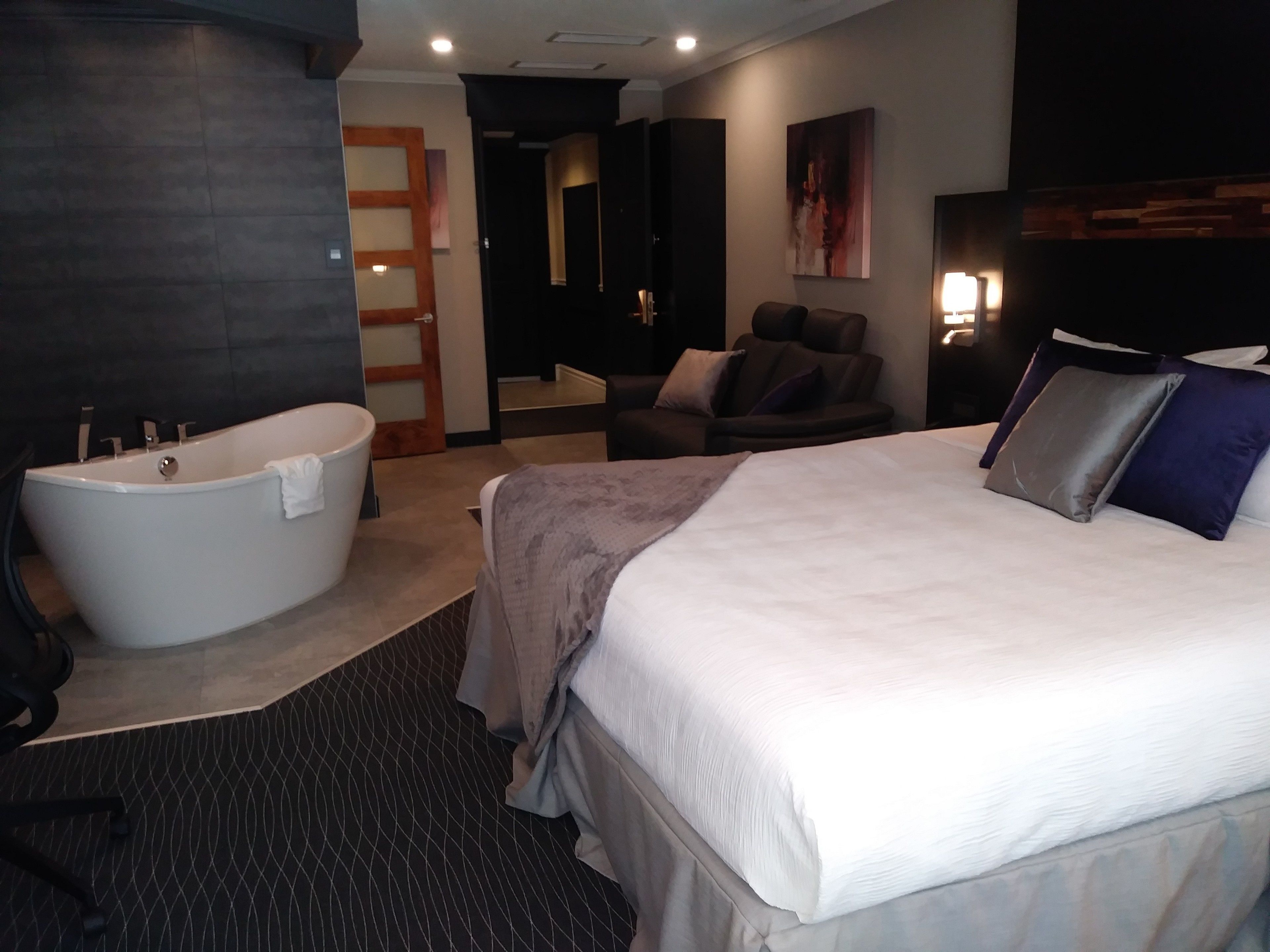 A modern hotel room with a large bed, a separate bathroom with a bathtub, a seating area with a chair, and a small table.