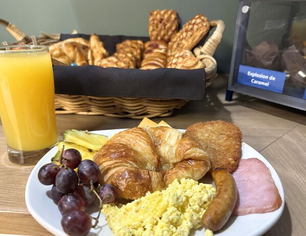 A breakfast plate with croissants, ham, scrambled eggs, sausage, and grapes, accompanied by a glass of orange juice and a basket of pastries. - Hôtel des Eskers (Amos)