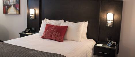 Habitación de hotel con una cama grande, ropa de cama blanca, un cojín rojo, lámparas de noche, una mesita de noche con un teléfono y un cuadro en la pared.