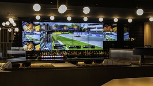 A bar area with multiple screens displaying a racing game, a bar counter with bottles, and seating. - Hôtel des Eskers (Amos)