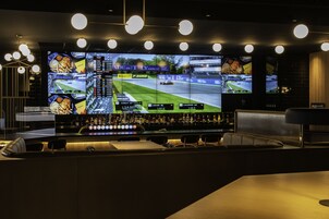 Zona de bar con varias pantallas mostrando un juego de carreras, barra con botellas y asientos.