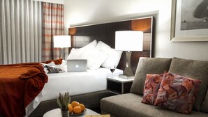 Quarto de hotel com cama, sofá, mesa de cabeceira com luminária, laptop, tigela de laranjas e quadro na parede.