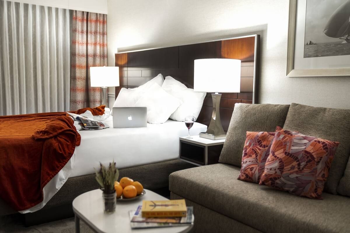 A hotel room with a bed, a sofa, a nightstand with a lamp, a laptop, a bowl of oranges, and a framed picture on the wall.