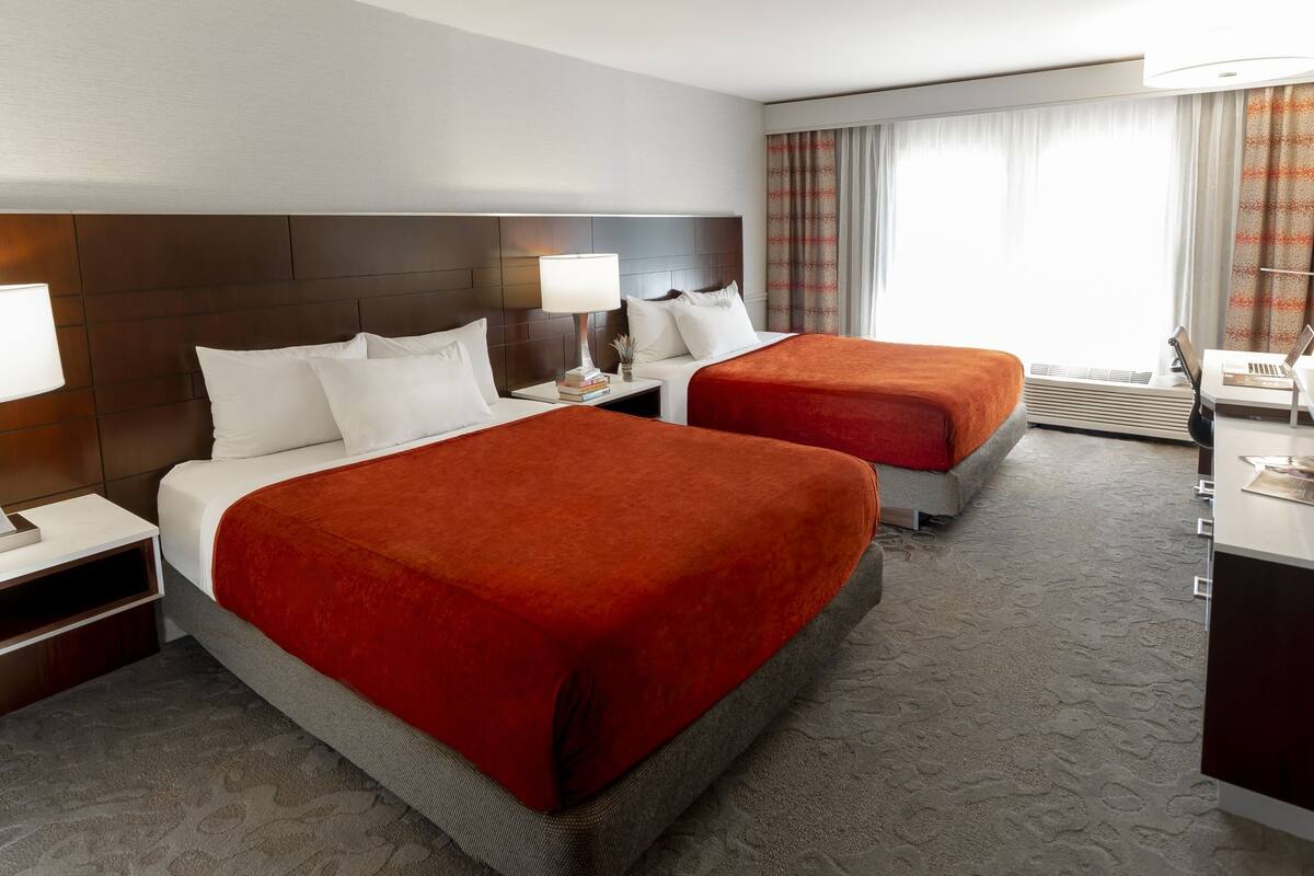 A hotel room with two beds, a desk, a TV, and a window with curtains.