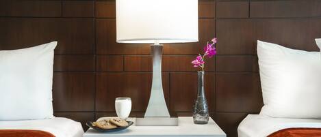 A hotel room with two beds, a nightstand with a lamp, a vase with flowers, a plate of cookies, and a glass of milk.