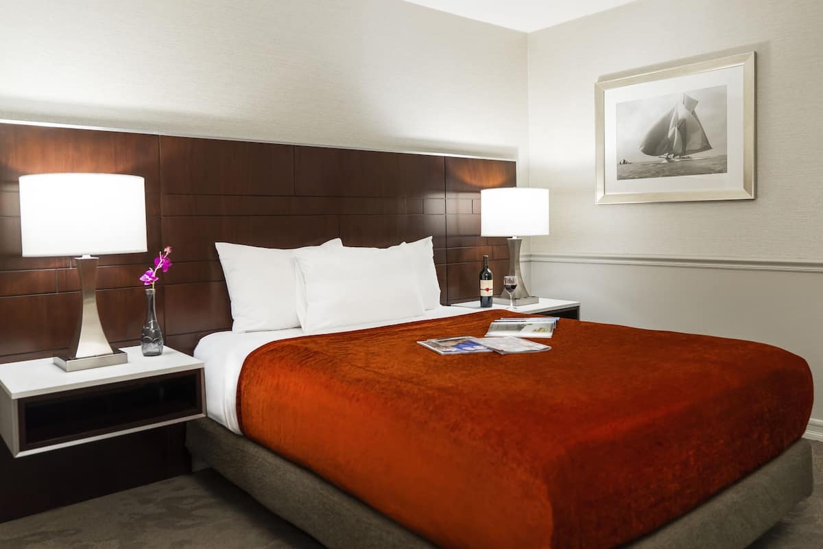 A hotel room with a large bed, two bedside tables, a nightstand, a framed picture on the wall, and a window with a view of the sea.
